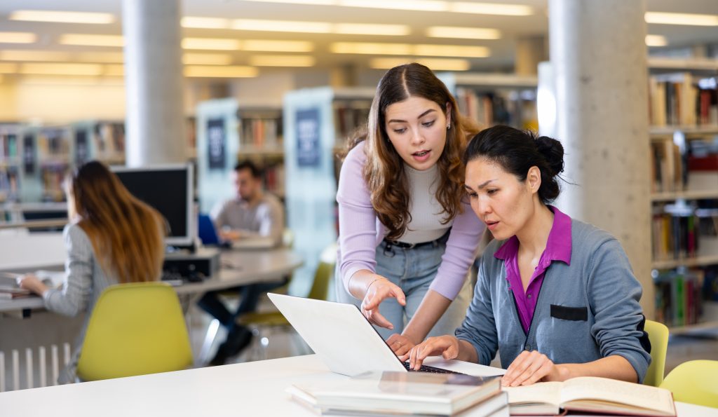 A student looking over the shoulder of a student explaining something on a computer in a library